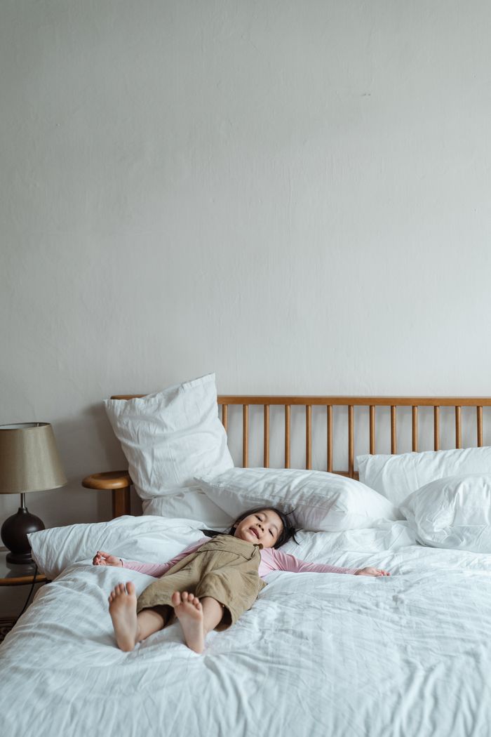 Young girl smiling while lying down comfortably on a bed with white linens, enjoying a relaxed moment indoors.