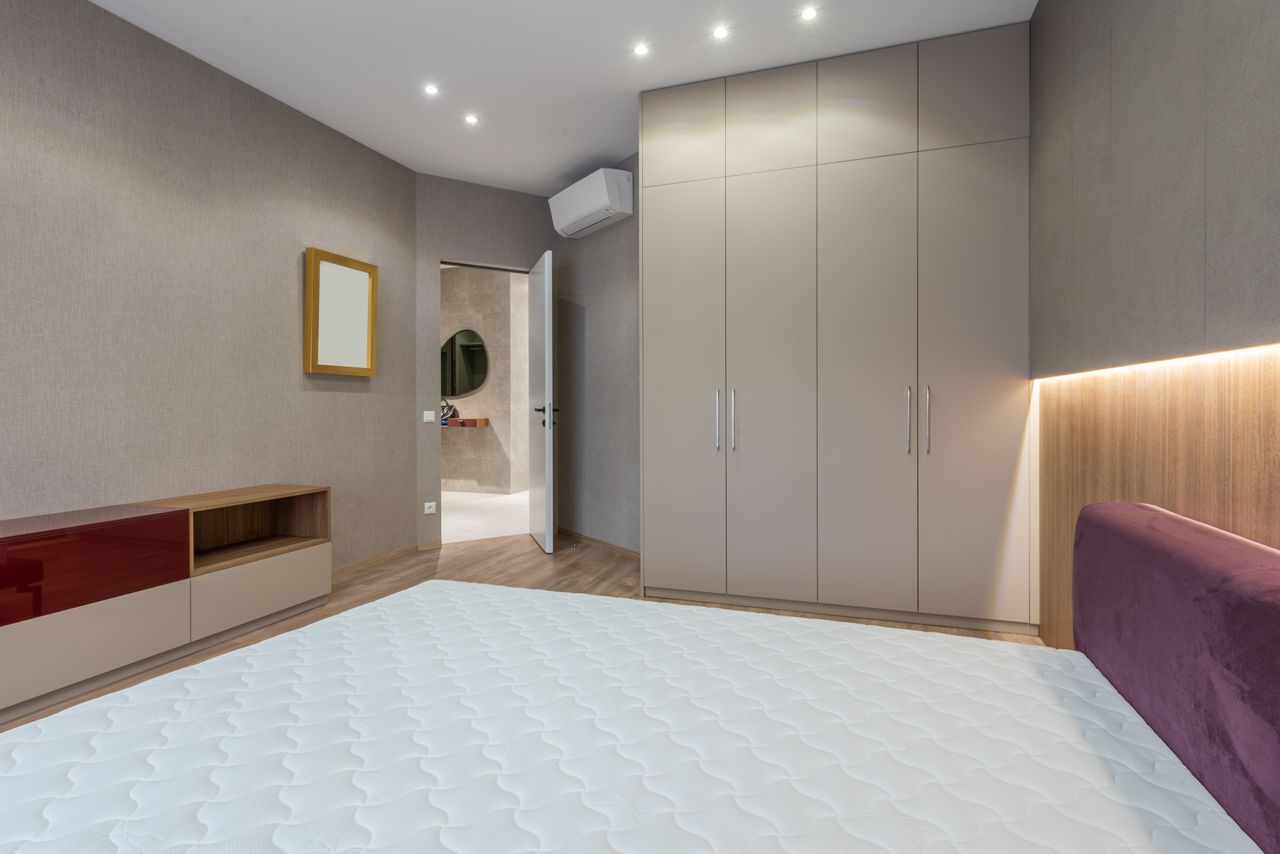 about-us-01 Interior of modern bedroom with white mattress on bed placed against cabinet near big closet and opened door in spacious apartment