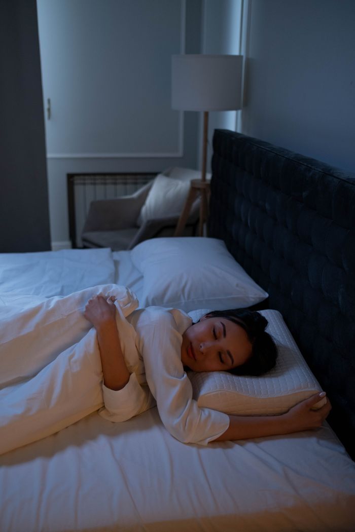 about-us-02 Asian woman peacefully sleeping in a cozy, dimly lit bedroom on white bedding.